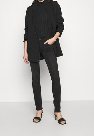 Femme portant un blazer noir croisé, un haut noir transparent texturé, un jean noir ajusté et des sandales à talons noires, debout avec une main dans la poche.
