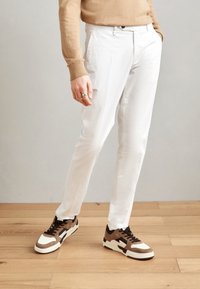 Pantaloni slim-fit bianchi abbinati a sneakers marroni e crema. Tessuto morbido con una finitura liscia, con chiusura classica a zip e bottone.