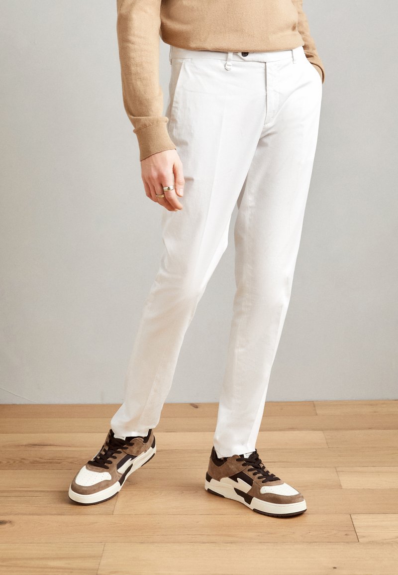 Pantaloni slim-fit bianchi abbinati a sneakers marroni e crema. Tessuto morbido con una finitura liscia, con chiusura classica a zip e bottone.