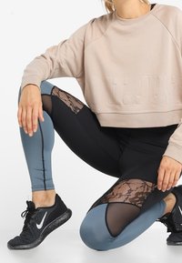 Felpa beige accorciata con testo in rilievo. Leggings neri e blu con pannelli in pizzo. Scarpe sportive nere con suole testurizzate.