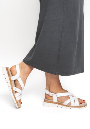 Weiße Sandalen mit einer Kork- und Gummisohle, mit gekreuzten Riemen und einer verstellbaren Schnalle. Getragen zu einem langen grauen Rock.