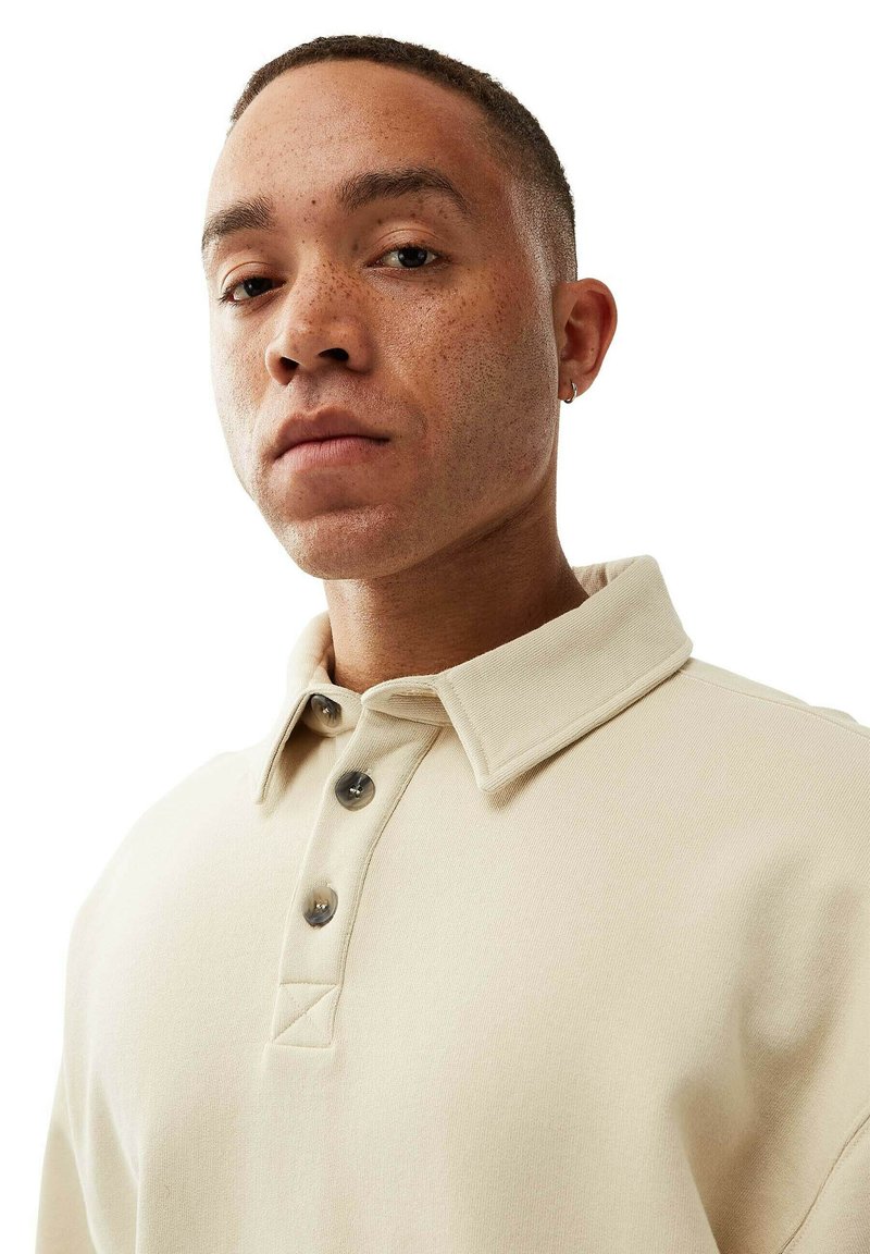 Beige polo shirt with a large collar, three button placket, and textured fabric. Close-up view highlights stitching details and fit.