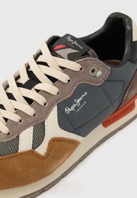 Hall ja pruun vabaajajalats seemisnahast ja nahast võrgusilma detailidega, beežide paelte ja küljel oleva logoga. Punane ja hall bränding.