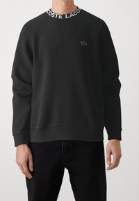 Černá mikina se žebrovaným límcem s nápisem "LACOSTE" v bílé barvě. Texturovaná tkanina, volný střih, malé logo Lacoste na prsou.