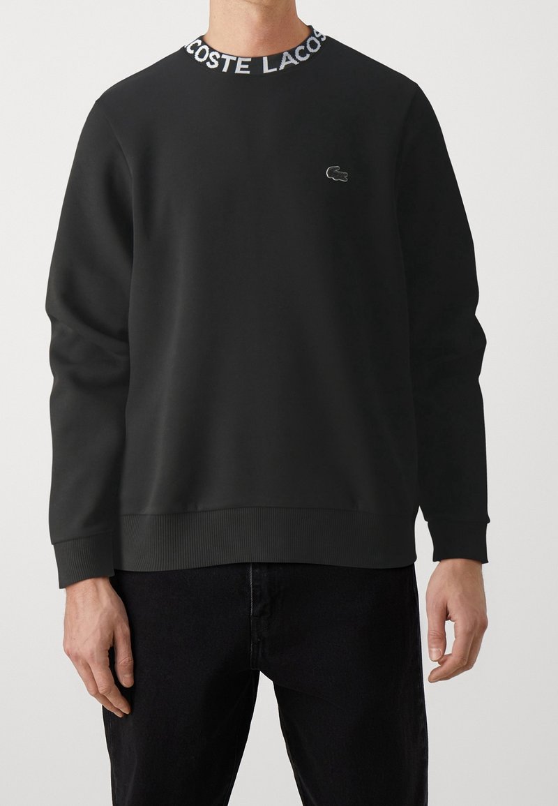 Černá mikina se žebrovaným límcem s nápisem "LACOSTE" v bílé barvě. Texturovaná tkanina, volný střih, malé logo Lacoste na prsou.