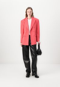 Koraalroze dubbelzijdige blazer met notch-revers, gecombineerd met zwarte faux-leer wijde broek. Model houdt een kleine zwarte handtas vast.