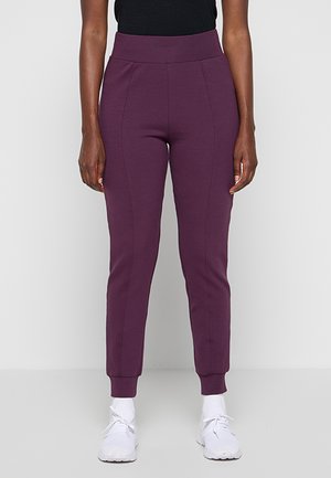 Femme portant un jogging slim taille haute violet avec des poignets côtelés, associé à des chaussettes blanches et des baskets blanches, debout sur fond blanc.