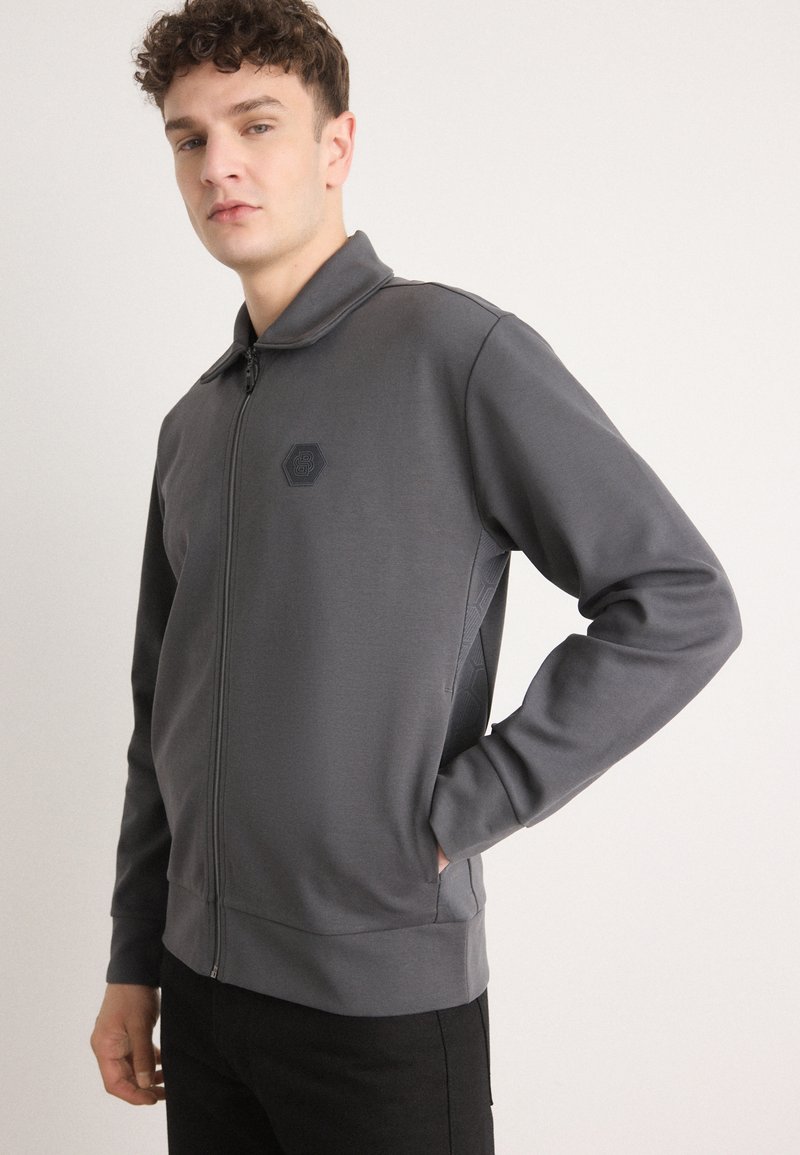 Giacca grigia con zip, caratterizzata da una finitura testurizzata, colletto alto e logo esagonale sul lato sinistro del petto, indossata con pantaloni neri.