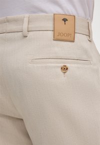 Beżowe bawełniane spodnie z skórzaną naklejką z logo "JOOP!", tylną kieszenią z guzikiem oraz teksturowaną powierzchnią materiału.