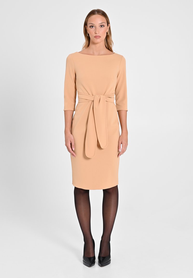 Knie-langes, hellbeiges Kleid mit Boots-Ausschnitt, dreiviertel Ärmeln und einem gebundenen Gürtel in der Taille; kombiniert mit schwarzen Strumpfhosen und Absätzen.