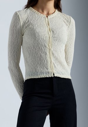 Kobieta ubrana w dopasowaną, kremową koronkową koszulę z długim rękawem i guzikami, zestawioną z czarnymi spodniami z wysokim stanem na jednolitym tle.