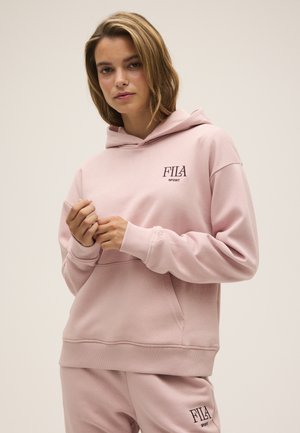 Kobieta ubrana w jasnoróżową bluzę z kapturem FILA Sport i pasujące spodnie dresowe, stojąca na tle jednolitego jasnego tła.