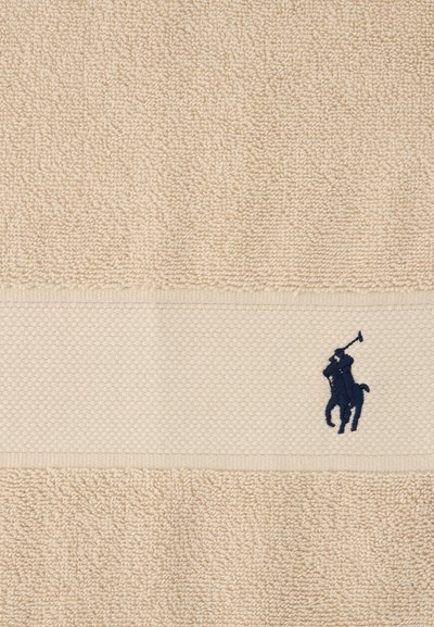 Πετσέτα μπεζ με υφή και υφαντή λωρίδα που έχει μικρή κεντημένη φιγούρα παίκτη πόλο σε σκούρο μπλε χρώμα που κρατάει μια μαλέτα.