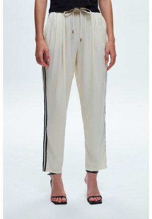 Femme portant un pantalon crème fuselé avec des bandes latérales noires, taille à cordon, et des talons noirs ouverts à l'avant, debout devant un fond blanc.