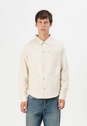Beige Hemdjacke mit Kragen, zwei Fronttaschen, Druckknöpfen und lockerer Passform. Aus strukturiertem Stoff, kombiniert mit blauen Jeans.