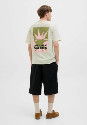 Lys beige t-shirt med grafisk design i grøn og pink med tekst, parret med mørke shorts og brune loafers.