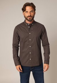 Bruine overhemd met knopen en een geometrisch patroon in rood en wit. Voorzien van een button-down kraag en lange mouwen, gecombineerd met donkerblauwe jeans.
