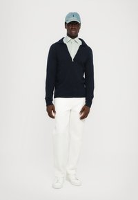 Suéter de cremallera de color navy, camisa de cuello azul claro, pantalones blancos y zapatillas blancas. La gorra es de color azul claro con un emblema. Tejido de punto de textura plana.