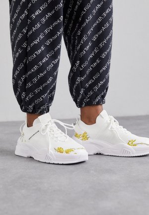 Białe sneakersy na sznurowadła z żółtymi barokowymi kwiatowymi wzorami, noszone z czarnymi spodniami z białym napisem "Versace Jeans Couture" drukowanym ukośnie.