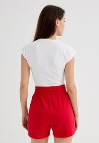 Top blanco de manga corta con mangas tipo cap, combinado con pantalones cortos elásticos de cintura roja brillante. El top es sencillo y los pantalones cortos son de longitud media, hasta el muslo.