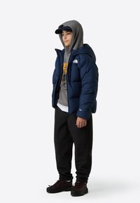 Sinine kapuutsiga pufferjakk, hall dressipluus julge kollase trükiga, mustad dressipüksid ja tumedad kingad. Vabaaja sportlik rõivakombinatsioon.