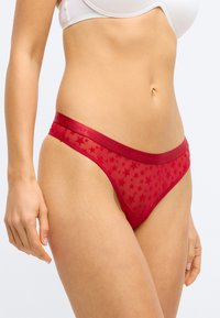 Rød mesh-tanga med stjernemønster, med en teksturert linning merket "TOMMY HILFIGER." Strømlinjeformet design med minimal dekning og gjennomsiktig stoff.