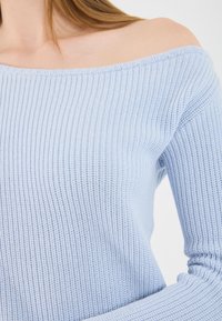 Primo piano di una donna che indossa un maglione a coste blu chiaro, off-the-shoulder, in cui si evidenziano dettagli del collo, della spalla e della parte superiore del braccio.