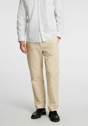 Chino chiari beige con una texture liscia, taglio dritto e tasche laterali, abbinati a una camicia bianca a righe con bottoni e scarpe nere.