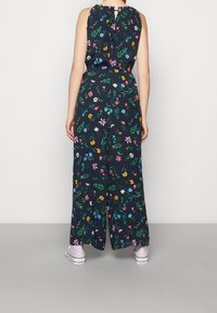 Blommig jumpsuit på mörk bakgrund med gröna blad; har ärmlös design, samlad halsringning, vida ben och rosa sneakers.