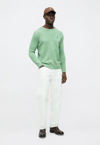 Zelený pletený sveter, biele nohavice, hnedá čiapka a čierne topánky. Sveter má textúrovaný vzor a rebrované manžety a lem.