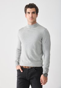Grauer, schmal geschnittener Rollkragenpullover aus weichem Strickstoff mit langen Ärmeln, kombiniert mit schwarzen Hosen und einem braunen Gürtel.