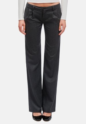 Donna che indossa pantaloni a gamba larga a righe grigio scuro con pieghe, abbinati a un top bianco a maniche lunghe corto e scarpe nere dalla punta affilata.