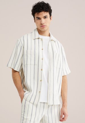 Jauns vīrietis valkā baltu īsroku krekli ar pogām un atbilstošas bikses ar plānām vertikālām melnām strīpām, pozējot vienkāršas fona priekšā.