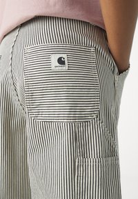 Gros plan de la poche arrière d'un pantalon rayé avec un écusson du logo Carhartt et la main d'une personne reposant dans la poche latérale, portant un haut rose.