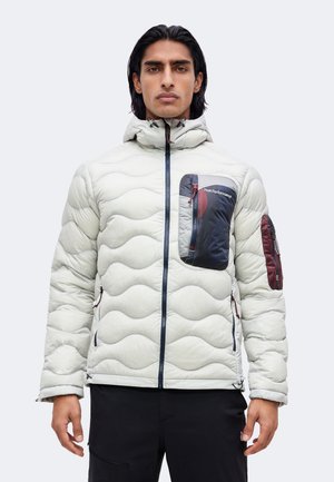 Homme portant une veste matelassée gris clair avec des poches zippées bleu marine et bordeaux sur la poitrine et les bras, debout devant un fond uni.