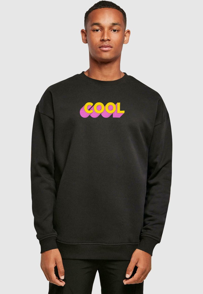 Mustmust must, mis on valmistatud puuvillast, kus on erksavärviline kollane ja roosa "COOL" graafika. Sisaldab ribatud varrukaotsasi ja alumist äärt ning on mugsalt lõigatud.