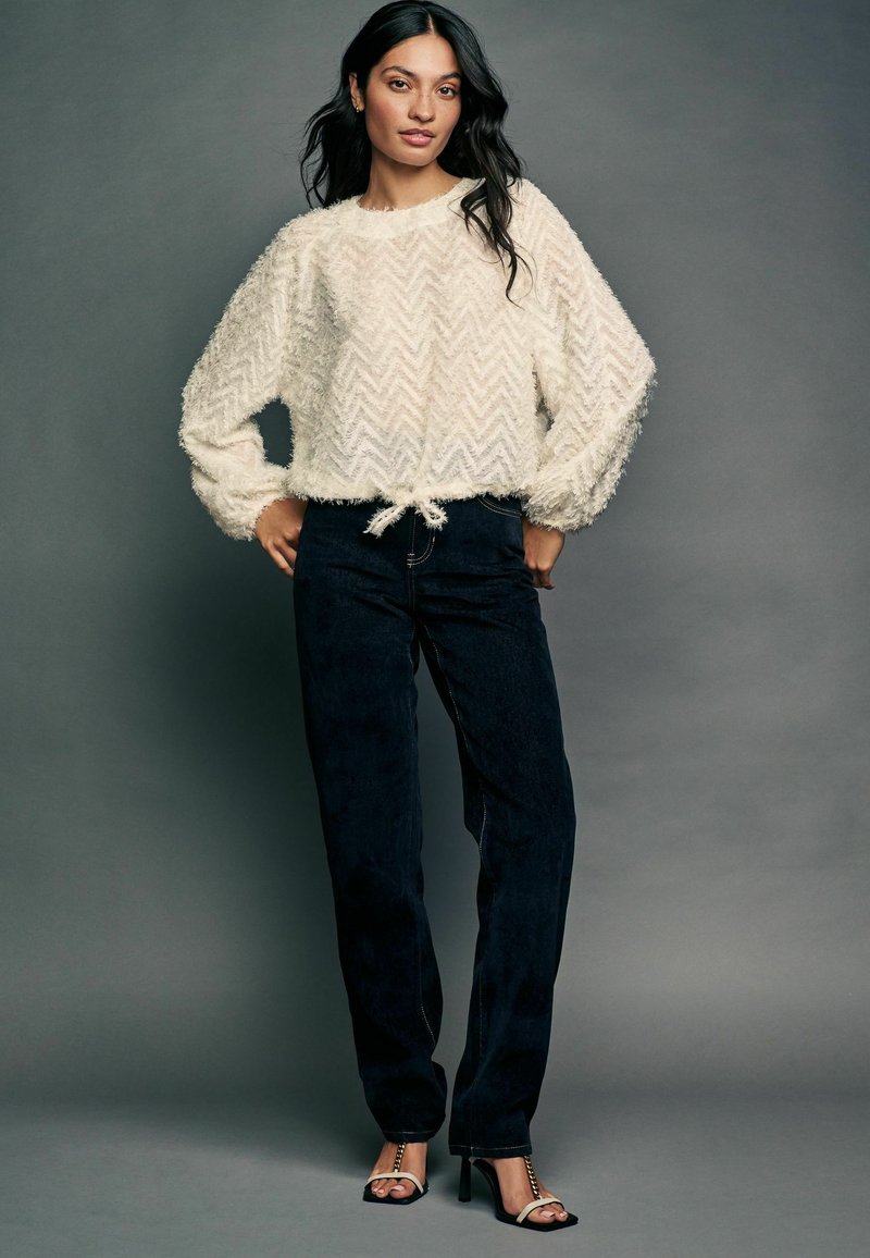 Ivory textured sweter z wzorem w zygzak i sznurkiem u dołu, w połączeniu z ciemnoniebieskimi spodniami dżinsowymi i czarnymi sandałkami na obcasie.