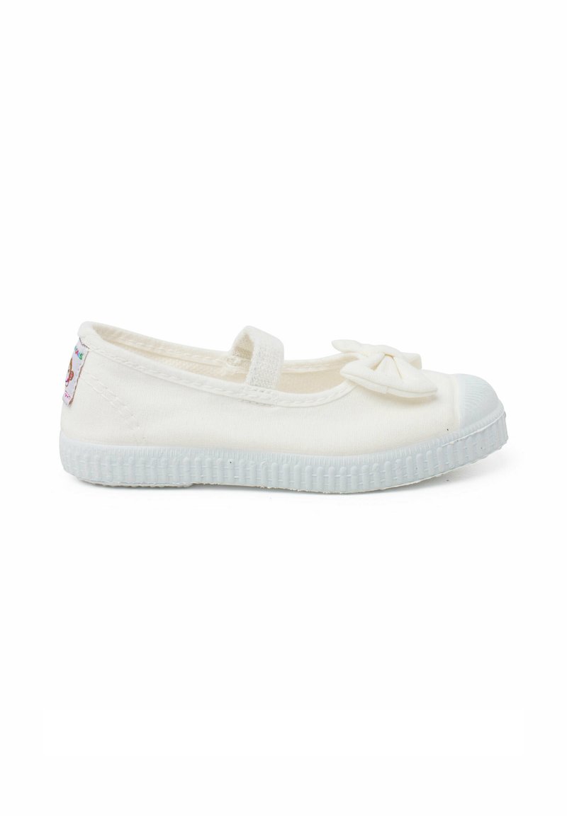 Zapato plano de lona blanca con puntera redonda, que presenta un pequeño lazo en la parte frontal y una suela de goma texturizada.