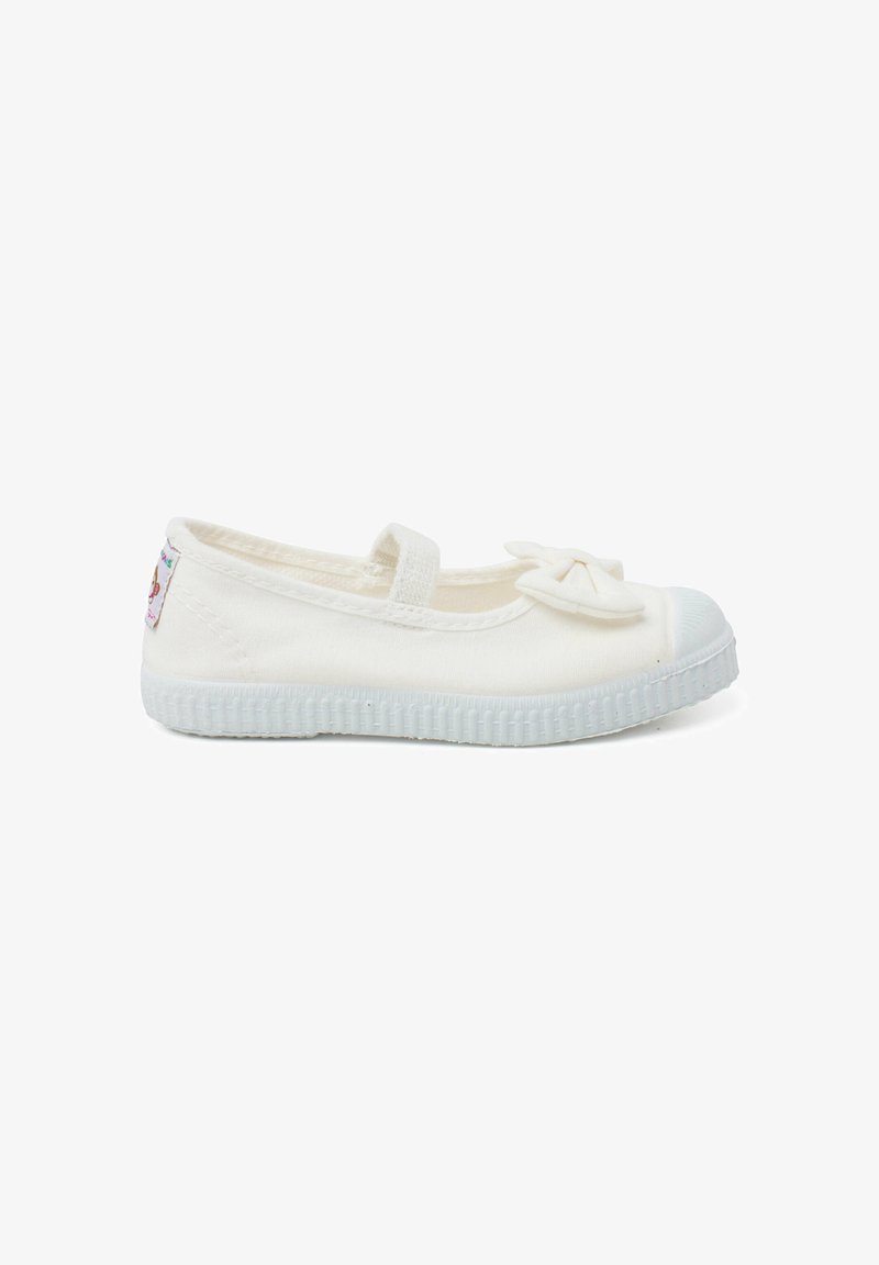 Zapato plano de lona blanca con puntera redonda, que presenta un pequeño lazo en la parte frontal y una suela de goma texturizada.