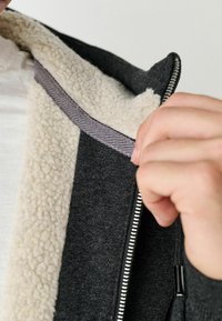 Dunkelgraue Jacke mit einem weißen, flauschigen Innenfutter. Das Innere hat einen gestreiften Stoffakzent; mit Reißverschluss und sichtbarem Handgriff.