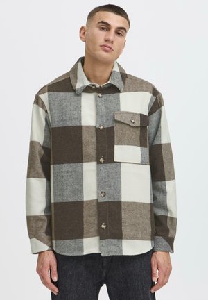 Ung mand iført en brun, grå og hvid ternet skjorte med knapper og brystlomme, kombineret med mørke bukser, stående foran en ensfarvet baggrund.