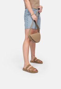 Tan schoudertas met een ronde vorm, een gladde leren textuur en een verstelbare schouderband. Gepaarde met denim shorts en bruine sandalen.