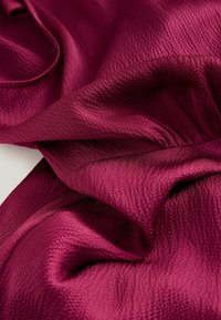 Tissu texturé bourgogne avec une surface légèrement froissée, drapé et superposé, mettant en valeur sa couleur riche et sa qualité tactile.