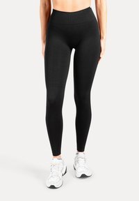 Leggings neri a vita alta realizzati in tessuto elasticizzato, con una texture liscia e una vestibilità aderente, abbinati a scarpe da ginnastica bianche.