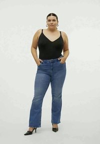 Mujer con una blusa sin mangas negra, jeans azules y tacones negros, de pie con las manos en los bolsillos contra un fondo claro y liso.