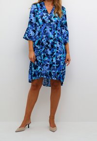 Kvinna klädd i en blå blommig knälång klänning med trekvartsärmar och beige spetsiga kitten heels mot en neutral bakgrund.