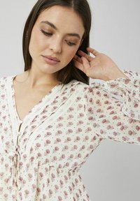 Blouse blanche à imprimé floral dans des tons roses. Caractérisée par un galon en dentelle, des détails de boutons et des poignets légèrement volantés. Tissu doux et léger.