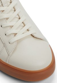 Zapatilla de cuero blanca con puntera redonda, superficie texturizada y cordones planos. Presenta una suela de goma de color marrón claro.