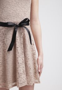 Robe en dentelle beige avec un ruban noir noué en nœud autour de la taille, montrant le bras et la main du modèle sur un fond uni.