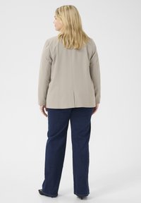 Blazer beige con maniche lunghe e spacco sul retro, abbinato a jeans a gamba dritta blu scuro. Tessuto liscio, dettagli minimi.